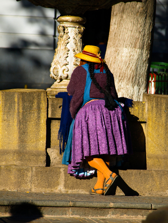 Bolivian woman - cholita - in typical colorful dress with violet skirt and yellow hut standing at fountain. View from back side. Potosi, Boliviaの写真素材