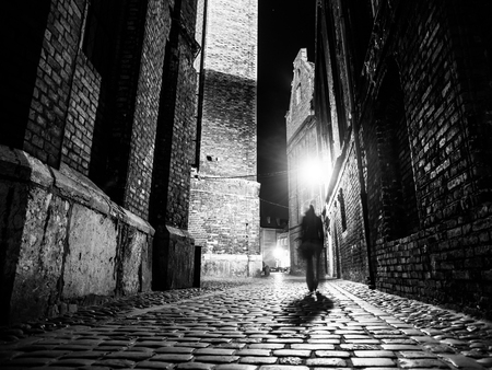 Illuminated cobbled street with light reflections on cobblestones in old historical city by night. Dark blurred silhouette of person evokes Jack the Ripper. Black and white image.の写真素材
