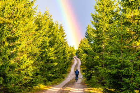 Mountain bike cycling after rain. Woman rides towards bright rainbow through forest country road. Outdoor sport theme.