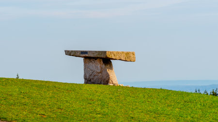 A solitary ancient rock table stands against a backdrop of clear blue sky atop a lush green hillside, with a calm sea visible in the distance.の写真素材