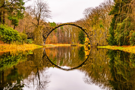 The Rakotzbrucke, also known as Devils Bridge, is reflected in calm waters on an overcast autumn day, surrounded by vibrant foliage. Germanyの写真素材