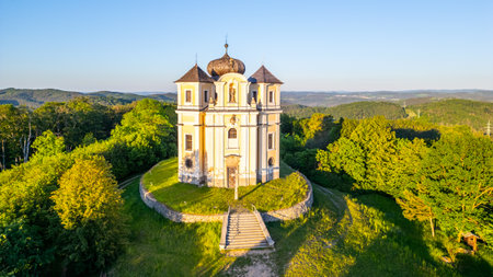 An aerial view of the Church of Saint John the Baptist and Our Lady of Mount Carmel at Makova hora, surrounded by lush green trees under a clear blue sky at sunset.の写真素材