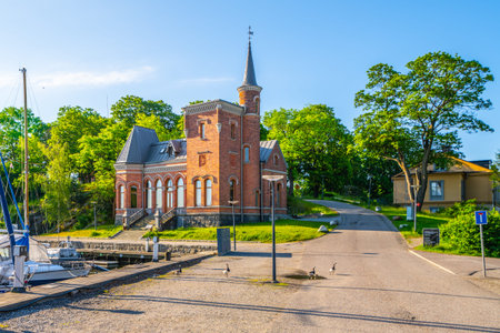 A serene summer day at Skridskopaviljongen, featuring a historic building surrounded by lush greenery and a clear blue sky, with boats moored nearby and ducks crossing the path. Stockholm, Swedenの写真素材