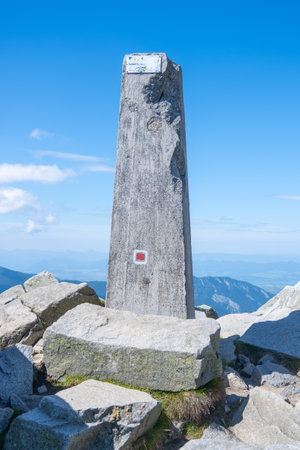 At the summit of Dumbier in the Low Tatras, a stone monument stands among rocky terrain, marking the highest point with a clear blue sky above and distant mountains in the background.の写真素材