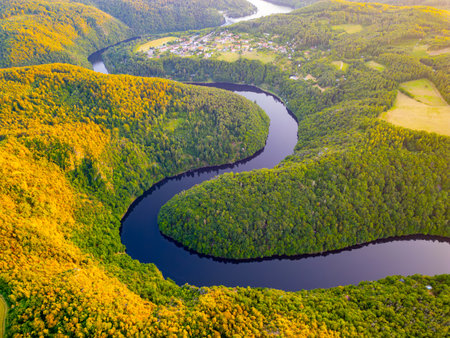 The vibrant sunset casts a warm glow over the winding Vltava River, surrounded by lush green hills and dense forests in Czechia, creating a serene and picturesque landscape.の写真素材