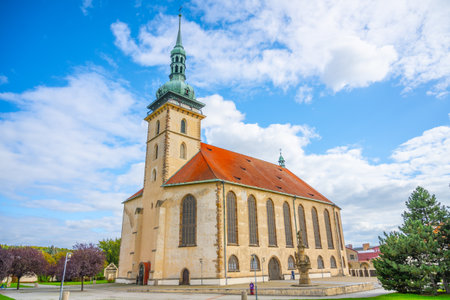 The Church of the Assumption of the Virgin Mary stands prominently in Most, Czechia, showcasing late Gothic architecture against a vibrant sky, surrounded by lush greenery.の写真素材