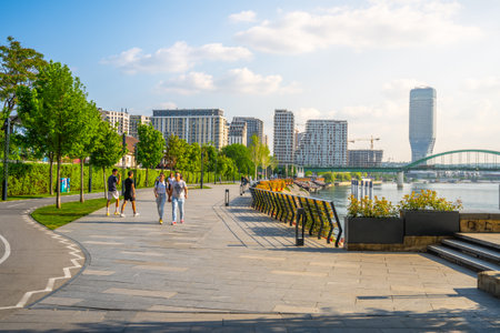 People stroll along the Sava Promenade in Belgrade, enjoying the greenery and scenic views of the river and modern architecture under a clear blue sky.の写真素材