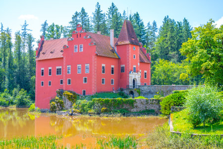 Cervena Lhota Castle gracefully rises from a rocky island, encircled by a serene lake. Lush greenery frames the vibrant structure under a clear blue sky.の写真素材