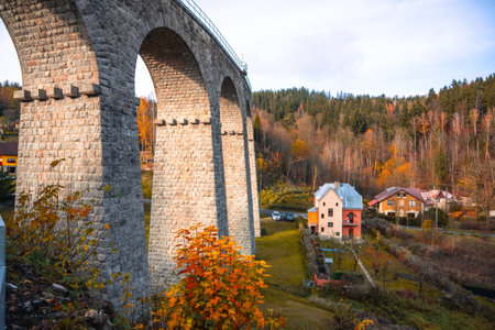 On a sunny autumn day, the impressive railway viaduct stands gracefully in Smrzovka, surrounded by vibrant fall colors and quaint houses under a clear blue sky.の写真素材