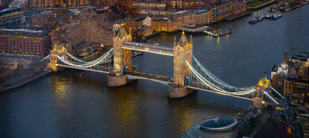 Tower Bridge stands majestically over the River Thames in London, glowing with lights as evening falls. Boats cruise beneath while the citys skyline sparkles with life.の写真素材