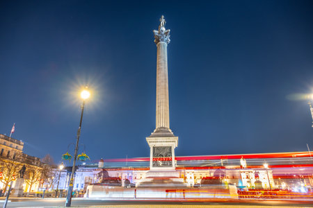 The majestic Nelsons Column stands tall in Trafalgar Square, beautifully illuminated against the night sky, surrounded by the lively atmosphere of London streets.の写真素材