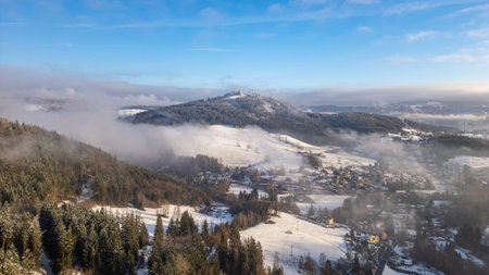 Aerial view captures the snowy terrain surrounding Tanvaldsky Spicak in Czechia. Fog drifts over hills while the village nestles beneath the snow-capped peaks under a clear sky.の写真素材