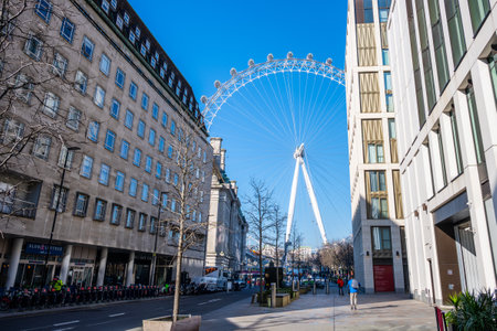 A clear blue sky provides a stunning backdrop to the London Eye towering over the street. Visitors stroll by modern buildings, enjoying the lively atmosphere of the city.の写真素材