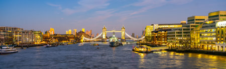Tower Bridge and HMS Belfast stand majestically over the Thames River during a tranquil evening in London, illuminating the skyline with warm lights as day turns to night.の写真素材