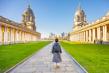 A visitor walks along the pathway amidst the stunning historical buildings of Greenwich Campus in London. With a clear blue sky, the city skyline can be spotted in the distance.の写真素材