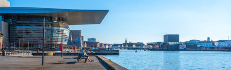 People relax by the water at the Copenhagen Opera House on a sunny day. The serene skyline and clear blue sky create a perfect atmosphere for leisure and enjoyment.の写真素材