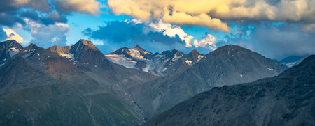 In the evening, the majestic alpine peaks are illuminated by soft light, casting dramatic shadows and highlighting the rugged terrain. Clouds swirl overhead, adding depth to the stunning summer landscape.の写真素材