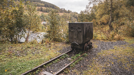 A mining truck sits on rusted tracks surrounded by trees and shrubs. The backdrop features rolling...