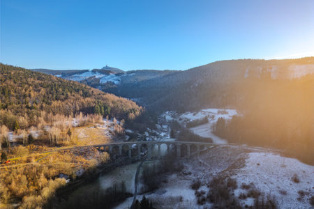 The railway stone bridge in Novina spans a tranquil valley, framed by snow-capped hills and Jested Mountain in the background during a beautiful winter sunset.の写真素材