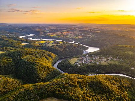 The Slapy Water Reservoir glimmers under a vibrant sunset, surrounded by lush hills and green fields. The winding Vltava River flows through the picturesque landscape, creating a tranquil ambiance.の写真素材