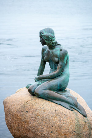 A bronze statue of a mermaid sits on a rock by the water in Copenhagen. This iconic landmark attracts visitors who admire its beauty and capture memories with its unique artistry.の写真素材