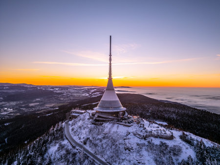 A breathtaking winter sunrise over Jested Mountain casts warm light across the snow-covered peaks and valleys. The distinctive tower stands tall against the colorful sky, creating a stunning contrast.の写真素材