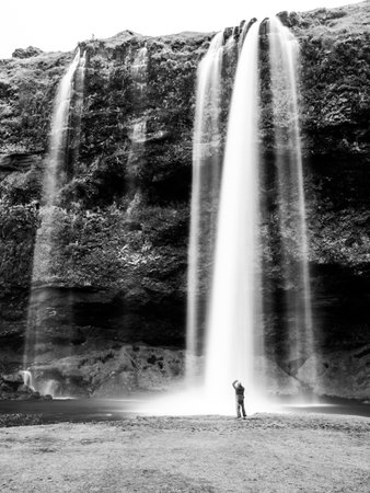 A lone individual stands in awe at the base of the cascading Seljalandsfoss Waterfall in Iceland, surrounded by the dramatic landscape. Black and white photography.の写真素材