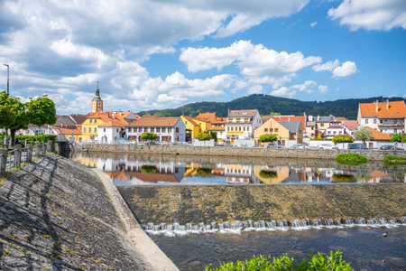 The tranquil waters of the Otava River reflect colorful buildings in Susice, Czechia. Visitors enjoy a sunny day with a backdrop of green hills and blue skies.の写真素材