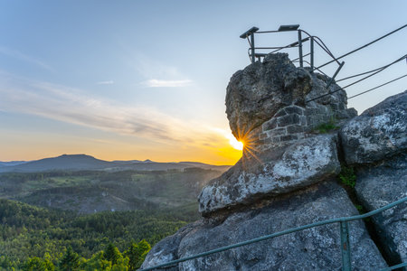 The sun sets behind Popov Cliffs, casting warm light over the sandstone rocks. Lush green forests stretch below, creating a stunning backdrop in Czechias Lusatian Mountains during the evening.の写真素材