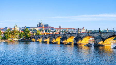 Prague Castle looms majestically in the background, while the historic Charles Bridge stretches over the Vltava River under a clear blue sky. A serene panorama captures the essence of Czechias charm.の写真素材