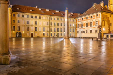 The Mrakotin Monolith stands tall in the courtyard of Prague Castle, surrounded by historic buildings. Soft lamp light reflects off the wet cobblestones, creating a serene nighttime atmosphere.の写真素材