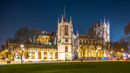 Illuminated Westminster Abbey stands tall against the night sky in London. Surrounding trees and lamp posts frame the majestic structure, showcasing its architectural beauty.の写真素材