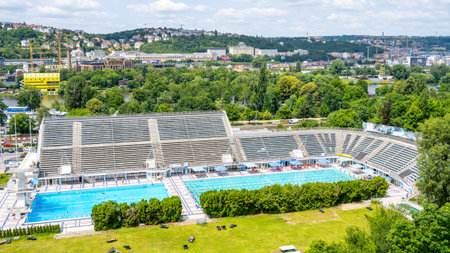 Visitors enjoy a sunny summer day at Podoli Swimming Pool in Prague. The outdoor pool area is bustling with people, surrounded by green trees and scenic views of the city.の写真素材
