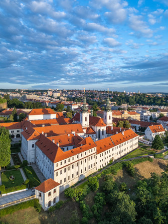 Aerial view of Strahov Monastery in Prague at sunrise. The golden light illuminates the red-tiled roofs and lush green surroundings, showcasing the historic architecture against a vibrant sky.の写真素材