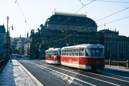 Two red trams travel across a bridge near the National Theatre in Prague on a sunny morning, showcasing the vibrant public transport system in this historic city.の写真素材