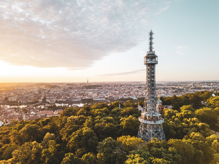 Golden light bathes Petrin Lookout Tower as the sun rises over Prague. Surrounded by lush greenery and the city skyline, this moment captures the beauty of early morning tranquility.の写真素材