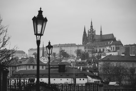 From a quiet street, the majestic Prague Castle looms in the background, framed by elegant lamp posts and the charm of historic rooftops. A captivating black and white view.の写真素材