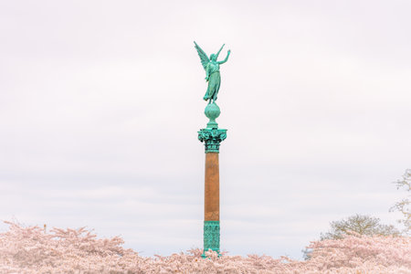 The Ivar Huitfeldt Column, a towering monument in Langelinie, Copenhagen, features an angelic figure atop a tall column, surrounded by pink cherry blossoms under a cloudy sky.の写真素材