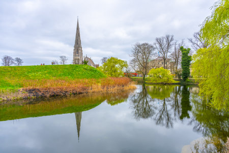 St Albans Church stands tall near Kastellet in Copenhagen, Denmark. Surrounding trees and grasses reflect in the calm water, creating a serene atmosphere on a cloudy day.の写真素材