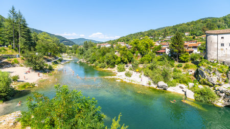 A tranquil stretch of Soca River meanders through Kanal, Slovenia, with green hills rising on either side. Nearby, people enjoy the clear water and warm sunshine, creating a lively atmosphere.の写真素材