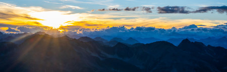 The sky lights up with hues of orange and gold as the sun sets behind the mountain range. Clouds drift lazily, creating a serene backdrop in this tranquil natural scene.の写真素材