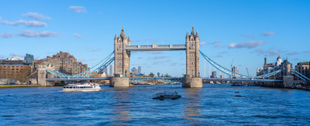 Tower Bridge stands majestically over the River Thames, showcasing its iconic architecture. A boat sails smoothly beneath the bridge on a bright and sunny day in London.の写真素材