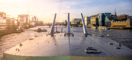 Turrets of HMS Belfast stand prominently along the River Thames, with the London skyline visible in the background during a peaceful sunset.の写真素材