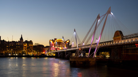 The illuminated Hungerford Bridge spans the River Thames in London during twilight. Surrounding lights reflect on the water, creating a peaceful evening atmosphere for viewers.の写真素材