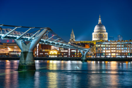 As evening falls, the Millenium Bridge elegantly spans the River Thames, showcasing stunning reflections while St. Pauls Cathedral stands majestically illuminated in the background.の写真素材
