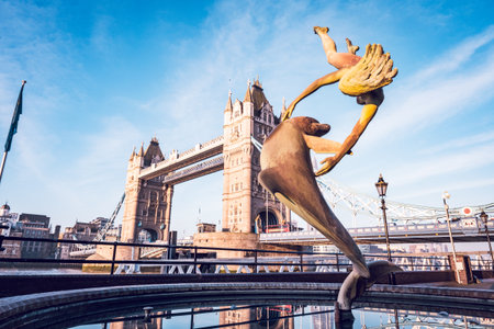 Girl with a Dolphin - vibrant statue of a girl joyfully interacting with a dolphin stands by the River Thames, showcasing the iconic Tower Bridge in the background under a clear blue sky.の写真素材