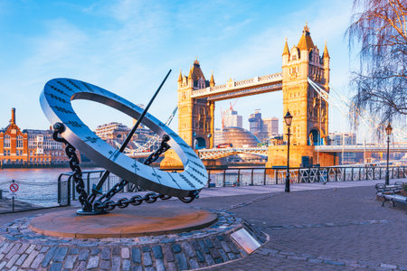 A large sundial stands prominently by the Thames River, with Tower Bridge in the background. The sun casts shadows on the sundial, marking the time while tourists enjoy the view.の写真素材