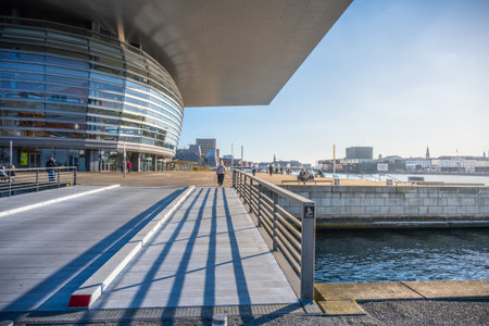 Visitors enjoy a bright sunny day at Copenhagen Opera House, taking in the stunning architecture and waterfront views. The vibrant atmosphere invites leisurely strolls along the canal.の写真素材