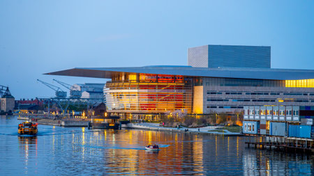 Copenhagen Opera House illuminates the evening sky with vibrant colors, reflecting beautifully on the water. Boats glide by as the cityscape provides a stunning backdrop in Denmark.の写真素材
