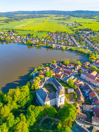 The castle in Rozmital pod Tremsinem stands proudly by the tranquil lake, surrounded by a picturesque town. Lush greenery and vibrant rooftops create a stunning view in the evening light.の写真素材